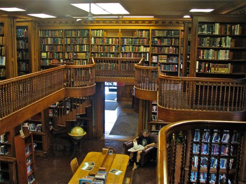 A cozy library with wooden shelves filled with books, spiral staircases, and a person reading at a table.