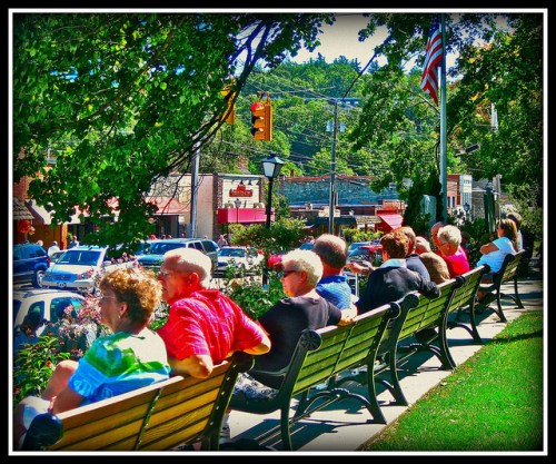 A row of people sitting on benches in a park, with shops and trees in the background on a sunny day.