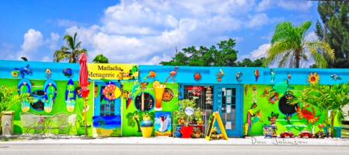 Colorful storefront with vibrant murals, tropical decorations, and lush greenery under a bright blue sky.