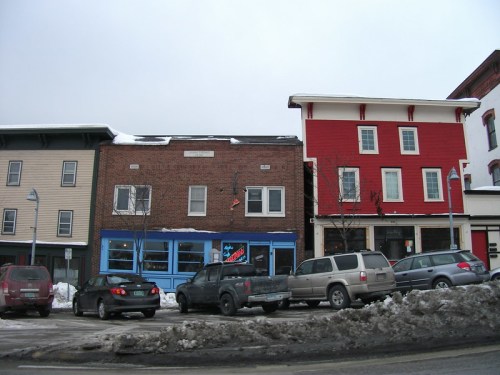 A snowy street scene featuring two buildings: one brick with blue accents and one red, alongside parked cars.