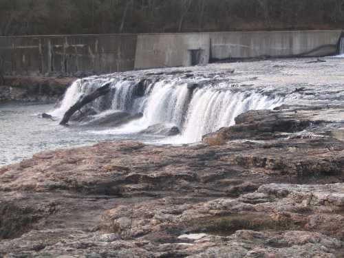 A waterfall cascading over rocky terrain, with a concrete structure in the background and trees lining the shore.