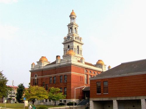 Historic building with a tall clock tower, orange domes, and red brick facade, surrounded by trees and a clear sky.