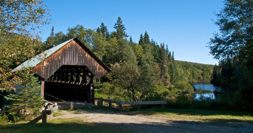 A scenic view of a covered bridge beside a calm river, surrounded by lush green trees and a clear blue sky.