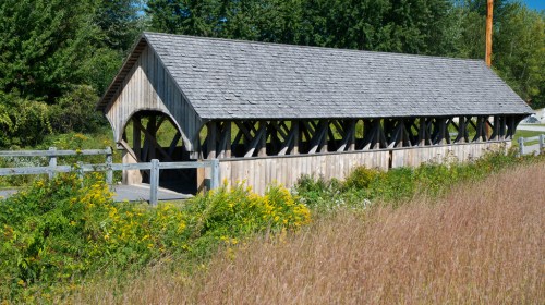 A wooden covered bridge surrounded by greenery and tall grass under a clear blue sky.
