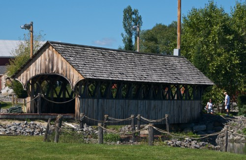 A rustic wooden covered bridge surrounded by greenery and a gravel path, with people walking nearby.