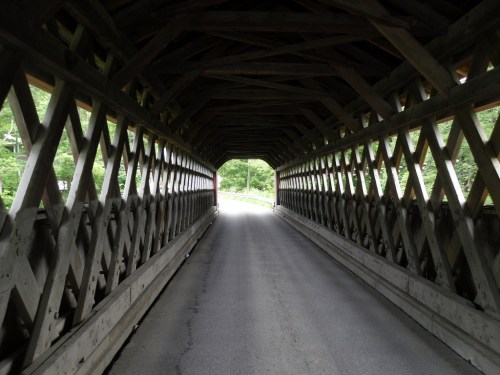 A view through a covered wooden bridge, showcasing a road leading to greenery beyond.