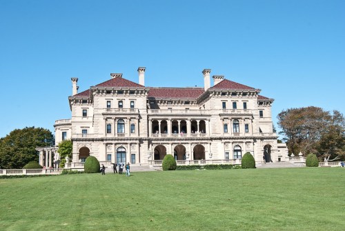 A grand mansion with a red-tiled roof, surrounded by green lawns and trees, under a clear blue sky.