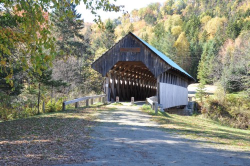 A rustic wooden covered bridge surrounded by colorful autumn trees and a gravel path leading to it.