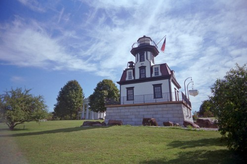 A white and black lighthouse stands on a grassy hill, surrounded by trees and under a blue sky with clouds.