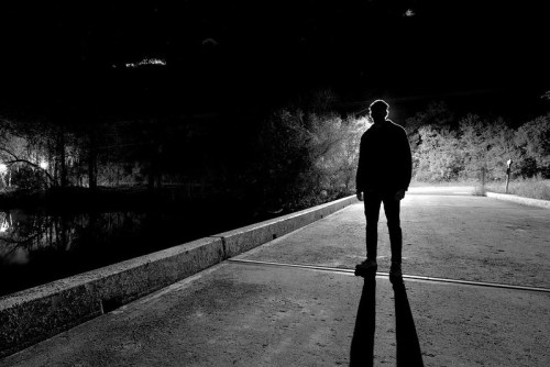 A silhouetted figure stands on a dark path by a body of water, surrounded by trees and faint lights in the background.