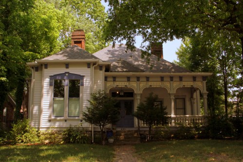 A charming vintage house with a porch, surrounded by lush green trees and a brick pathway.