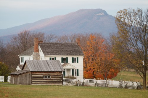 A white farmhouse and a rustic barn surrounded by autumn trees, with mountains in the background under a cloudy sky.