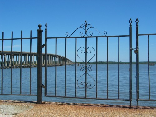A decorative black metal fence in front of a calm body of water, with a bridge visible in the background under a clear blue sky.