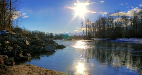 A serene river scene with sunlight reflecting on the water, surrounded by trees and rocky banks under a clear blue sky.