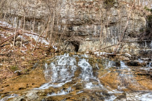 A serene waterfall cascades over rocky terrain, surrounded by trees and a rocky cliff in a tranquil natural setting.