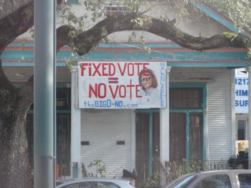 A large banner on a building reads "FIXED VOTE = NO VOTE" with a website link below it. Trees are in the foreground.