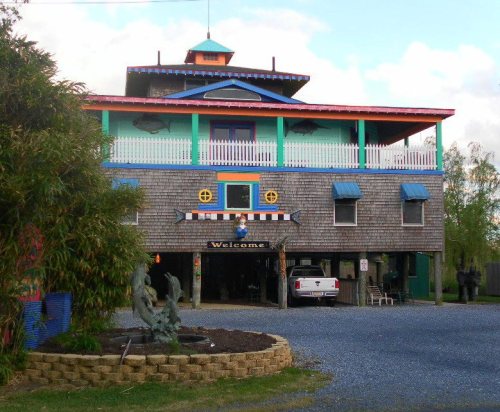 Colorful, multi-story building with a welcoming sign, surrounded by greenery and gravel parking area.