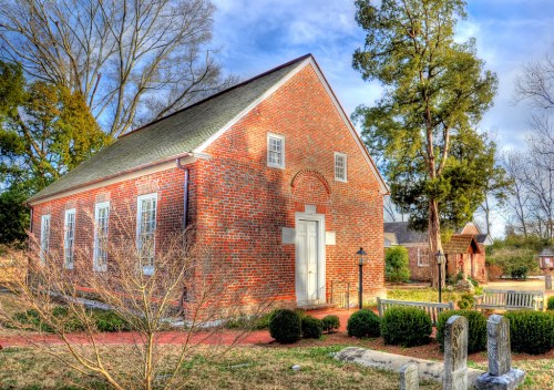 Historic brick building surrounded by greenery and trees, with a clear blue sky in the background.