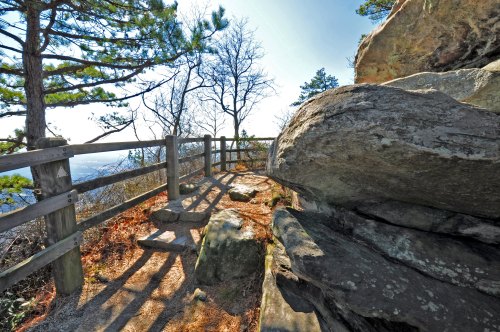 A rocky viewpoint with a wooden fence, surrounded by trees and a clear sky, offering a scenic landscape.