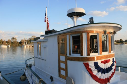 A white boat with wooden accents, featuring an American flag and patriotic bunting, docked on calm water.