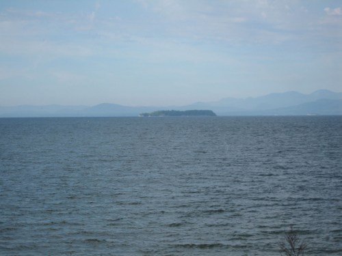 A serene view of a calm lake with a distant island and mountains under a blue sky.