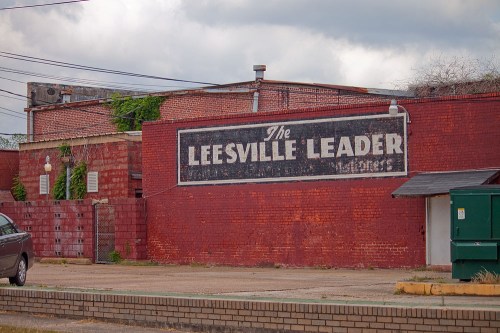 A weathered brick building with a faded sign reading "The Leesville Leader" against a cloudy sky.