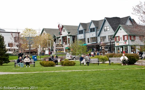 A park scene with people sitting on benches, surrounded by shops and trees on a sunny day.