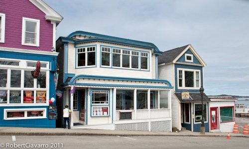 Colorful buildings line a street, featuring a blue-trimmed house and a small shop, with a cloudy sky above.