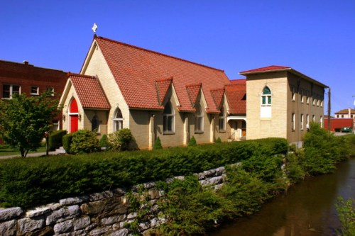 A brick church with a red roof and door, beside a small stream, surrounded by greenery and buildings in the background.