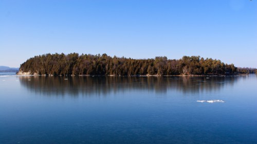 A serene island surrounded by calm water, with trees reflecting on the surface under a clear blue sky.