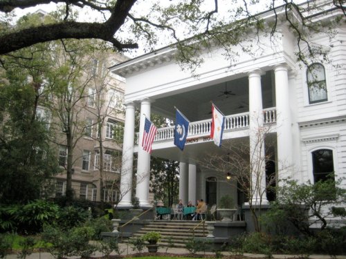A historic white house with columns, featuring flags and people sitting on the porch surrounded by greenery.