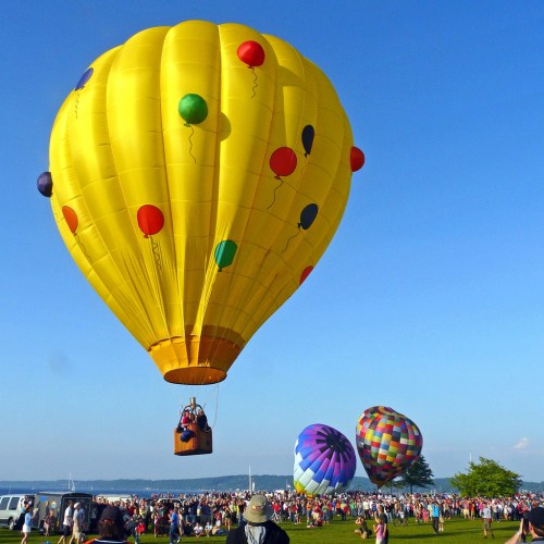 A vibrant yellow hot air balloon floats above a crowd at a festival, with colorful balloons in the background.