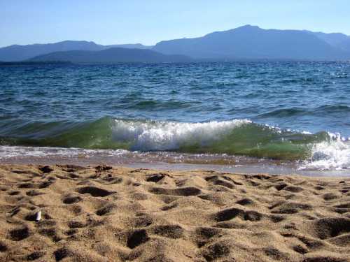 A sandy beach with gentle waves lapping at the shore, framed by mountains under a clear blue sky.