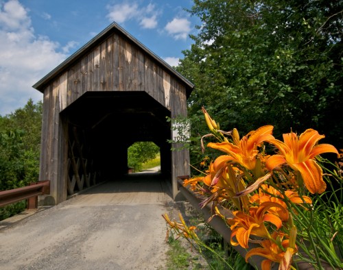 A wooden covered bridge with a gravel path, surrounded by lush greenery and vibrant orange flowers in the foreground.