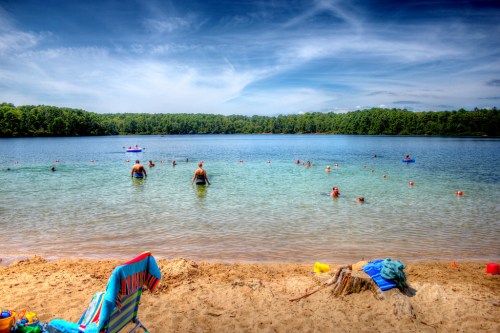A sandy beach with people swimming in clear water, surrounded by trees and a blue sky with clouds.