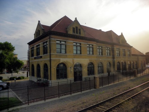 Historic train station building with a yellow facade and red roof, surrounded by a fence and railway tracks.