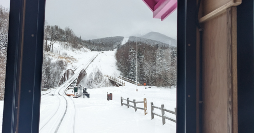 View from a train window showing snowy tracks, a tunnel, and a mountainous landscape in the background.