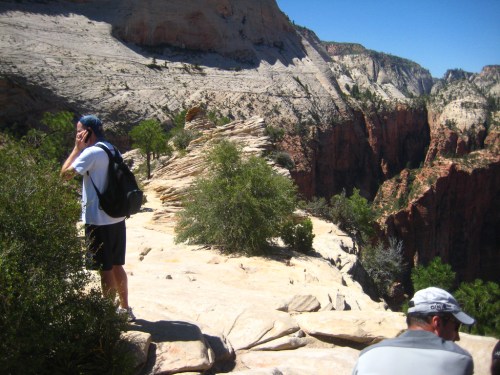 A hiker stands on a rocky ledge, overlooking a canyon with steep cliffs and greenery under a clear blue sky.