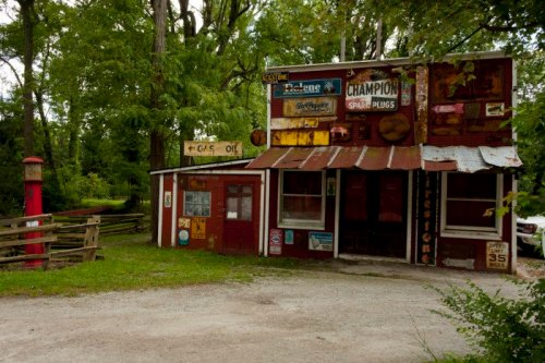 A rustic, weathered building with vintage signs, surrounded by trees and a gravel path.