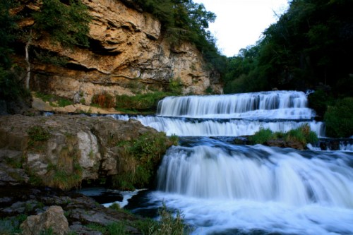 A serene waterfall cascades over rocky ledges, surrounded by lush greenery and rugged cliffs.
