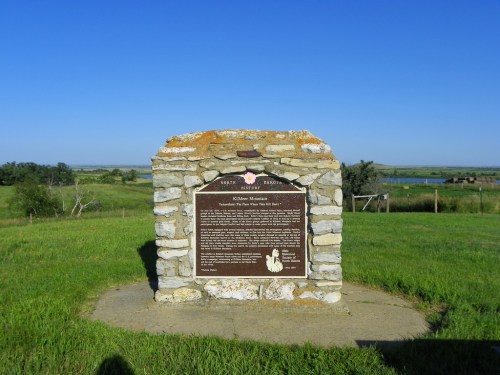 A stone monument with a plaque stands in a grassy area, overlooking a scenic landscape and blue sky.