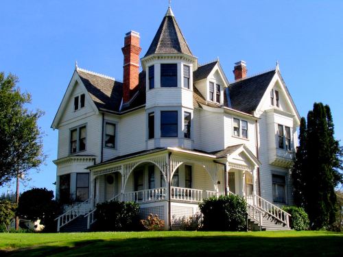 A large, elegant Victorian-style house with a turret, white siding, and a wraparound porch, set against a clear blue sky.