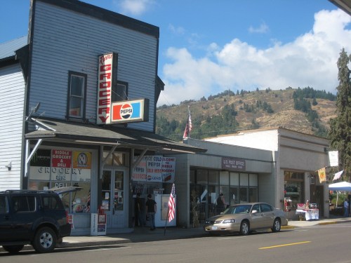 A small town street featuring a grocery store, post office, and mountains in the background under a blue sky.