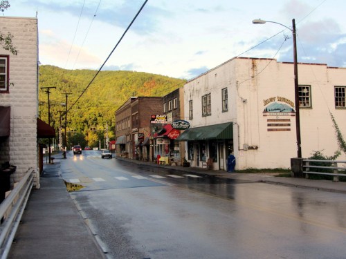 A quiet street lined with small shops and buildings, surrounded by green hills under a partly cloudy sky.