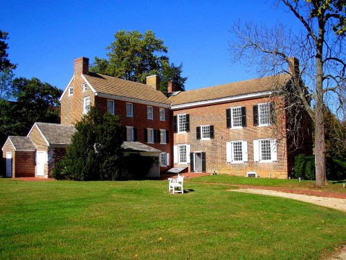 Historic brick house with a large lawn, surrounded by trees under a clear blue sky. A white chair is visible in the yard.