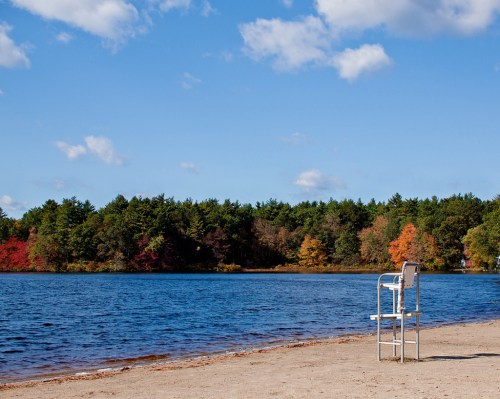 A lifeguard chair on a sandy beach by a calm lake, surrounded by trees with autumn foliage under a blue sky.