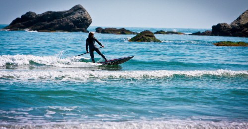 A person in a wetsuit paddleboards on calm ocean waters with rocky formations in the background.