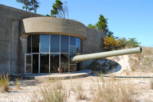 A concrete bunker with large glass windows, featuring a protruding tank barrel in a sandy landscape.
