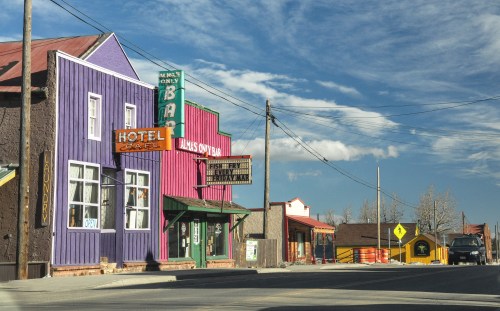 Colorful buildings line a quiet street, featuring a hotel and a bar under a clear blue sky.