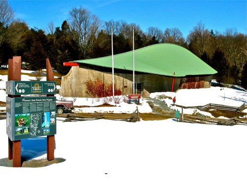 A modern building with a green roof, surrounded by snow, next to an informational sign and trees in the background.